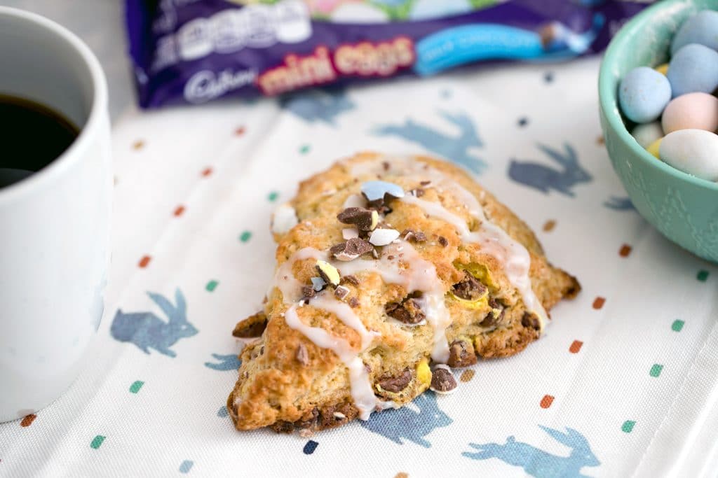 Landscape view of a Cadbury Mini Egg scone on a bunny towel with bag of mini eggs, bowl of mini eggs, and cup of coffee in the background