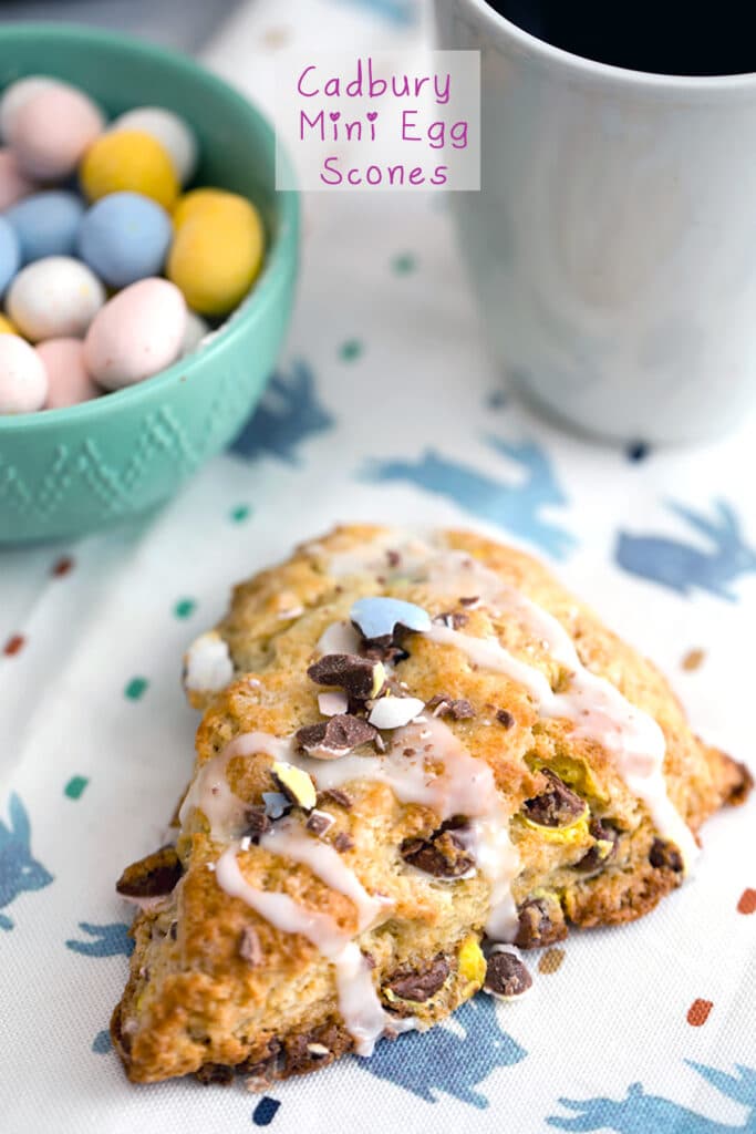 Overhead view of a Cadbury Mini Egg scone on a bunny towel with a bowl of mini eggs and a cup of coffee in the background and recipe title at top.