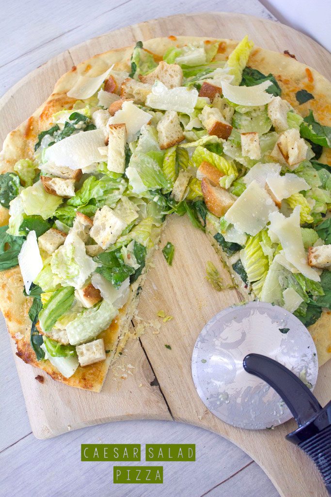 Overhead view of Caesar Salad Pizza with a slice removed on a pizza paddle with pizza cutter and recipe title at bottom