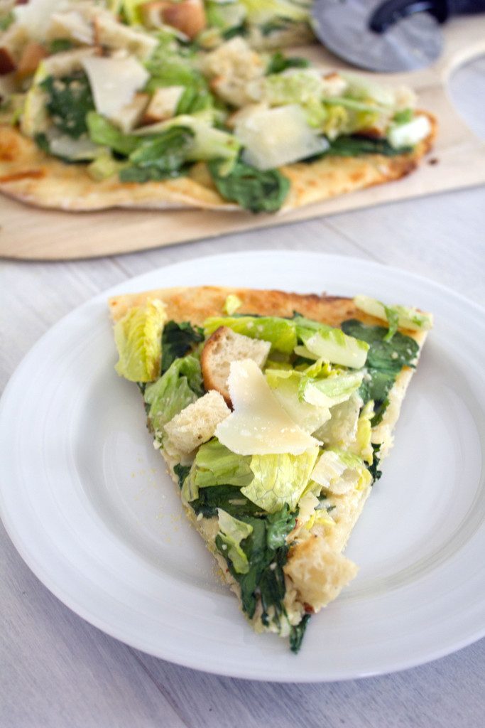 Overhead close-up view of a slice of Caesar salad pizza on a white plate with rest of pizza and pizza cutter in background