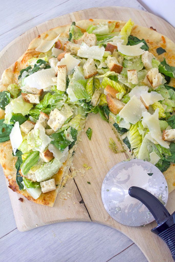 Overhead view of Caesar Salad Pizza with a slice removed on a pizza paddle with pizza cutter