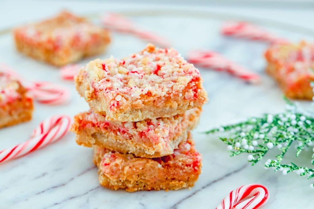 Landscape head-on view of three candy cane crumble bars stacked on top of each other with more candy cane bars in the background, along with mini candy canes and holly