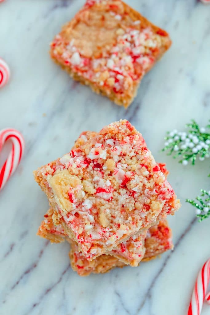 Overhead view of three candy cane crumble bars stacked on top of each other with another bar behind them, all on a marble surface with mini candy canes and holly