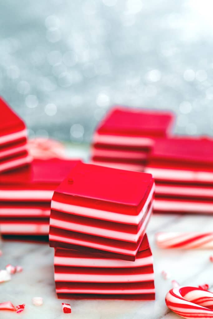 Head-on view of two red and white layered candy cane jello shots stacked on top of each other with crushed candy canes, mini candy canes, and more jello shots stacked in background.