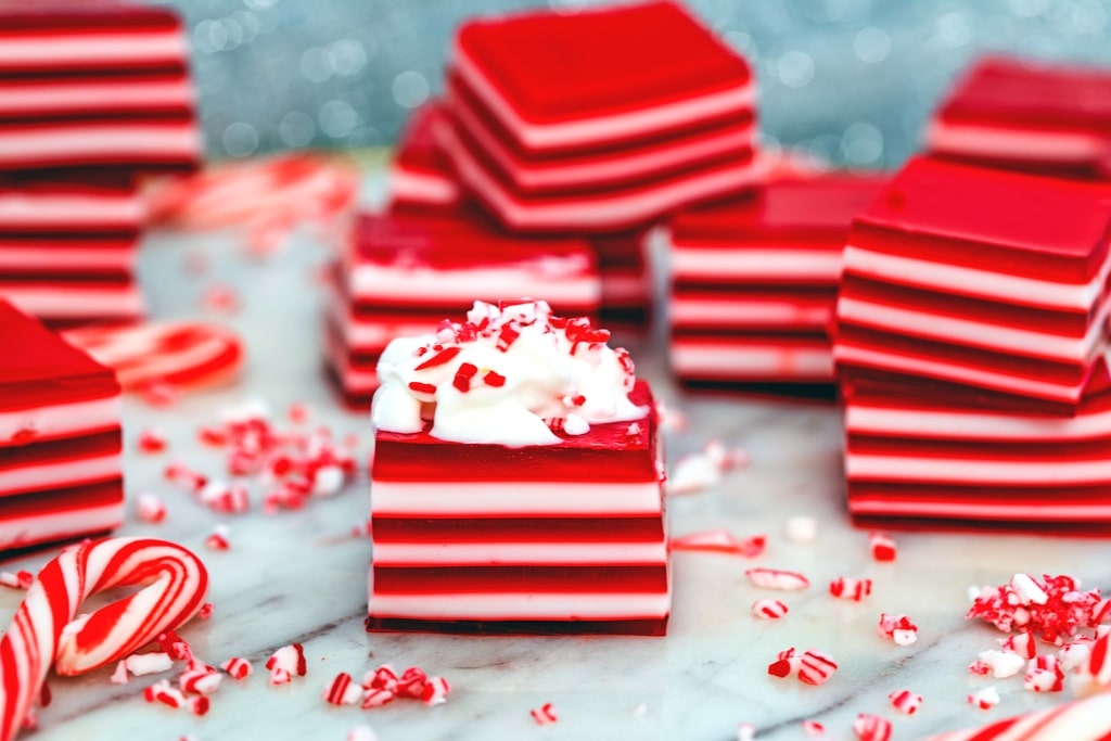 Landscape head-on view of multiple red and white layered candy cane jello shots, one with whipped cream on top with crushed candy canes and mini candy canes scattered around.
