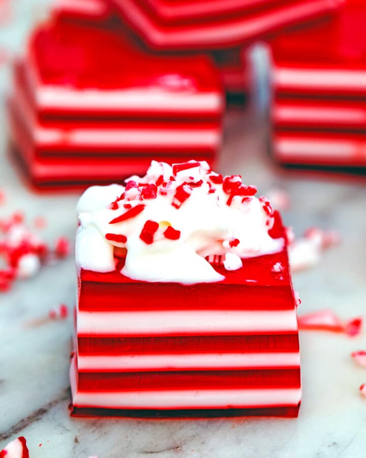 Head-on closeup view of a red and white layered candy cane jello shot topped with whipped cream and crushed candy canes with more jello shots in the background.