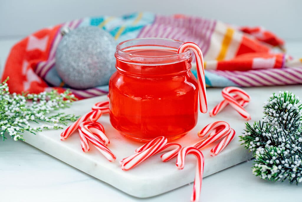 Landscape photo of a small jar of candy cane syrup with mini candy canes, ornaments, and holly all around.