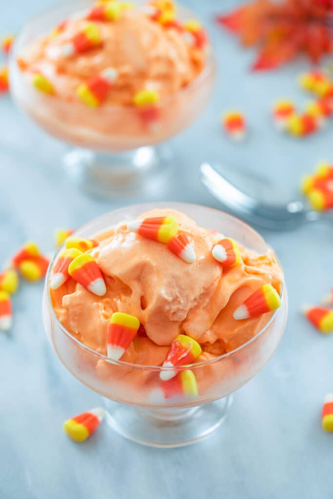 Overhead view of clear bowl of candy corn ice cream surrounded by candy corn, a spoon, and second bowl of ice cream in the background