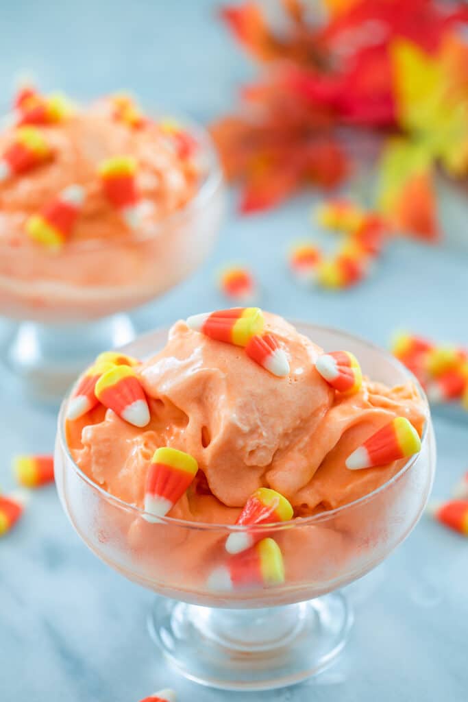 Close-up view of clear dish filled with candy corn ice cream topped with candy corn with more candies scattered around and second bowl of ice cream and leaves in the background