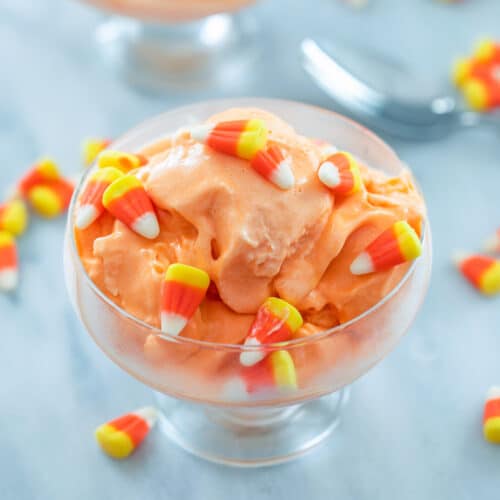 closeup view of clear bowl of candy corn ice cream surrounded by candy corn, a spoon, and second bowl of ice cream in the background