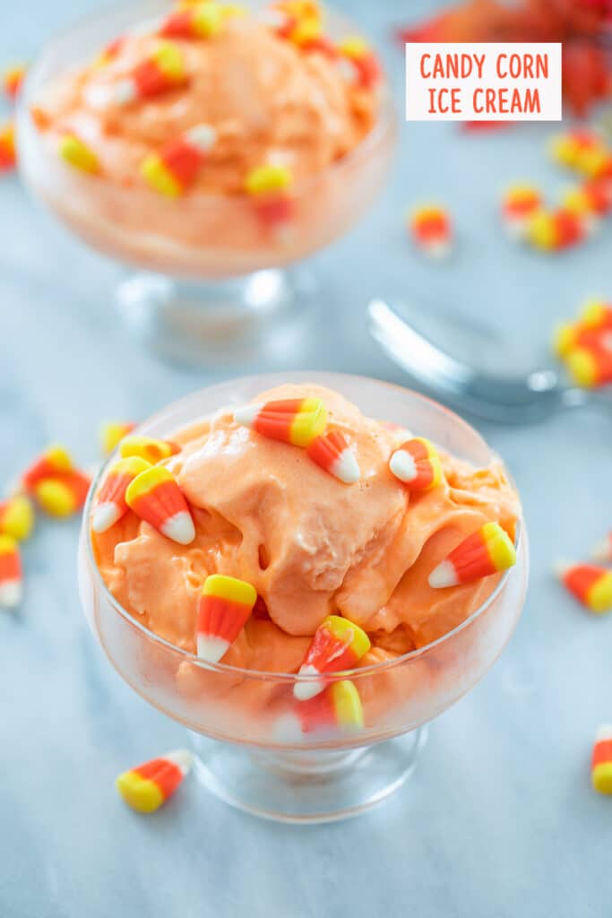 Overhead view of clear bowl of candy corn ice cream surrounded by candy corn, a spoon, and second bowl of ice cream in the background with "Candy Corn Ice Cream" text at top