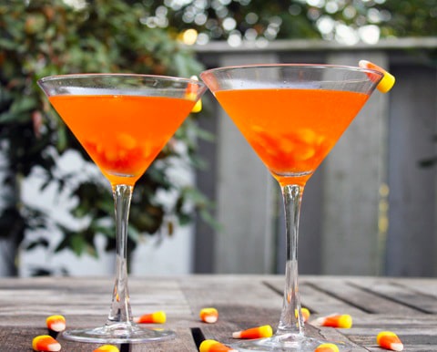 Head-on view of two bright orange candy corn martinis on a wooden table outside, surrounded by candy corn