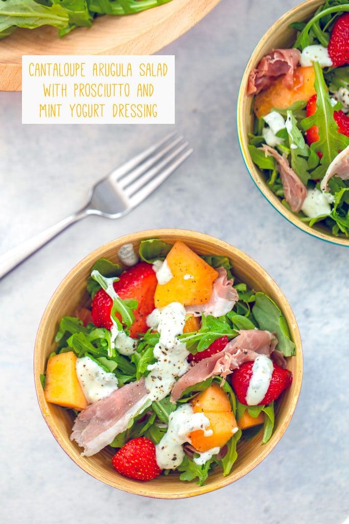 Overhead view of two bowls of Cantaloupe and Prosciutto Arugula Salad with strawberries and mint yogurt dressing drizzled over the top, fork in the background, and text at top