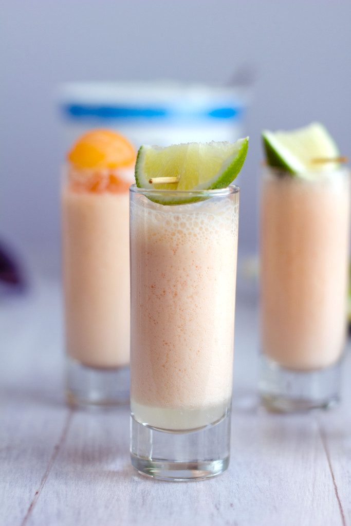 Head-on closeup view of a cantaloupe boozy smoothie shooter with lime garnish with more smoothie shooters in the background