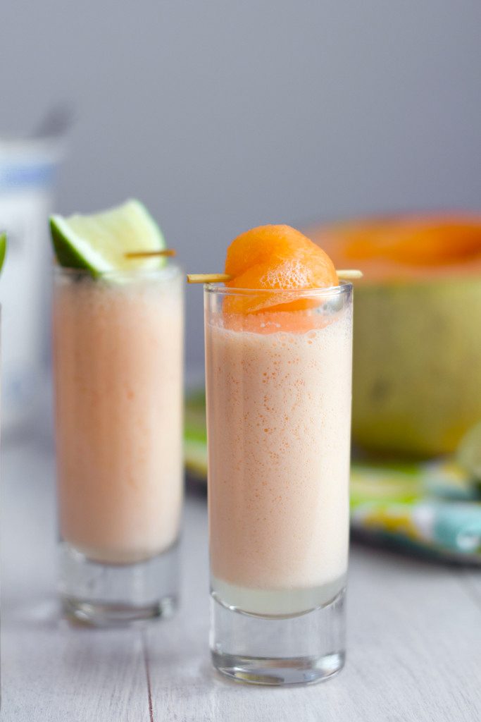 Head-on view of a cantaloupe boozy smoothie shooter with cantaloupe ball garnish with second smoothie with lime garnish and cantaloupe half in background