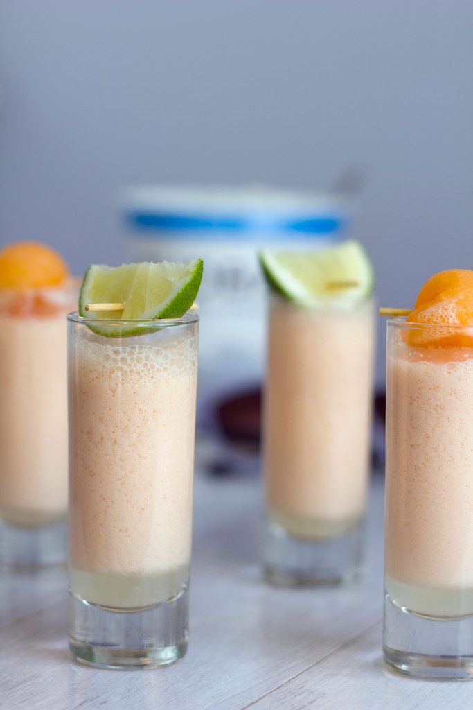 Head-on view of multiple cantaloupe boozy smoothie shooters with lime and cantaloupe garnishes with container of Greek yogurt in the background