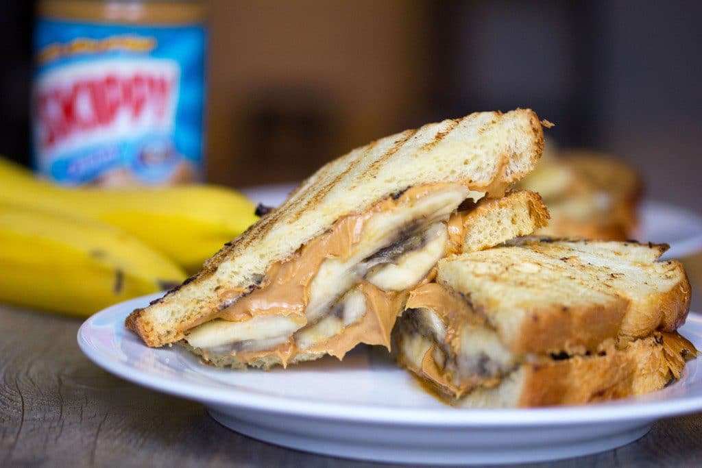 Landscape view of caramelized banana and peanut butter sandwich sliced in half on a white plate with bananas and peanut butter jar in background