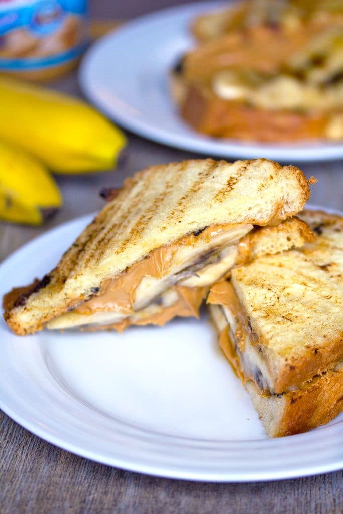 Head-on view of a caramelized banana and peanut butter sandwich cut in half on a white plate with a second sandwich, bananas, and peanut butter jar in the background