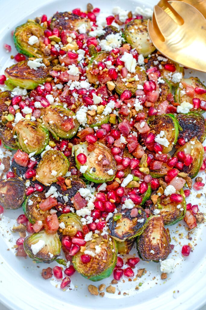 Overhead view of caramelized brussels sprouts with pancetta, pomegranate, and feta on a white platter with gold serving pieces