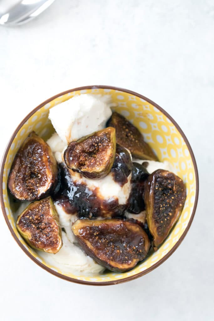 Bird's eye view of a yellow bowl filled with vanilla ice cream, caramelized fogs and their juices, with a spoon in the background