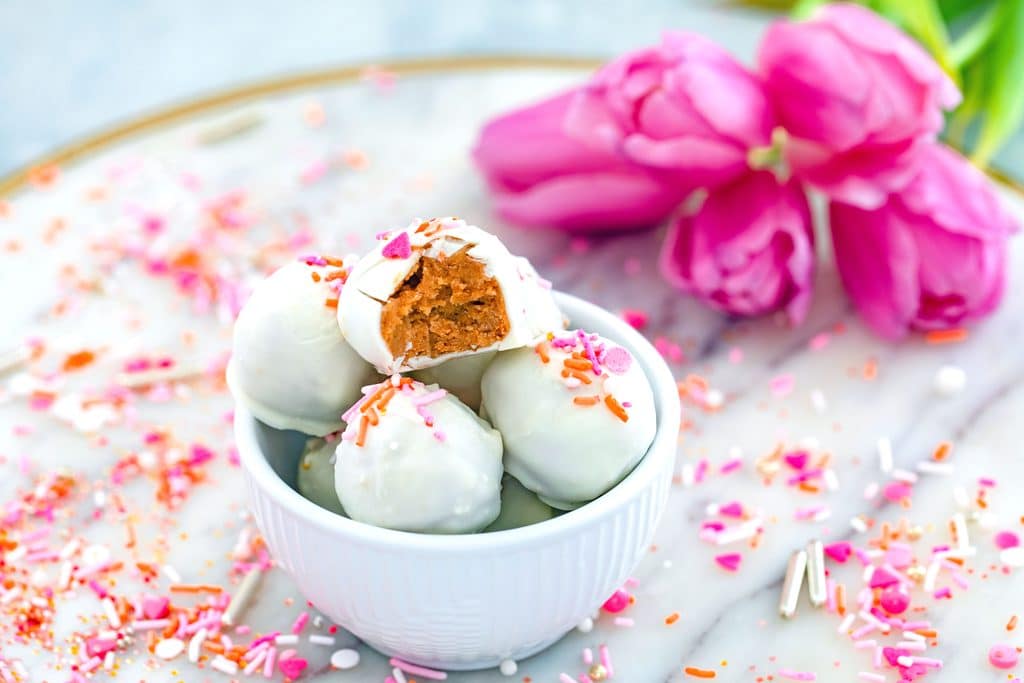 Landscape view of Carrot Cake Oreo truffles with a bite taken out of the top one  in a white bowl on a marble surface. Sprinkles are surrounding the bowl and pink tulips are in the background