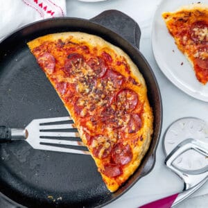 Overhead closeup view of half a cast iron skillet pizza with slices on plates on the side, pizza cutter, and red pepper flakes.