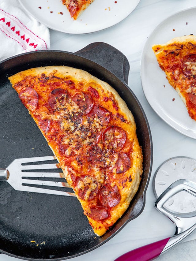 Pizza in a cast iron skillet with spatula lifting it, slice on a plate, and pizza cutter in background.