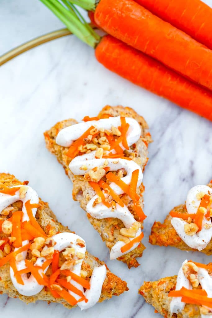 Overhead view of chai carrot cake scones drizzled with cream cheese frosting on a marble surface with bunch of carrots in the background