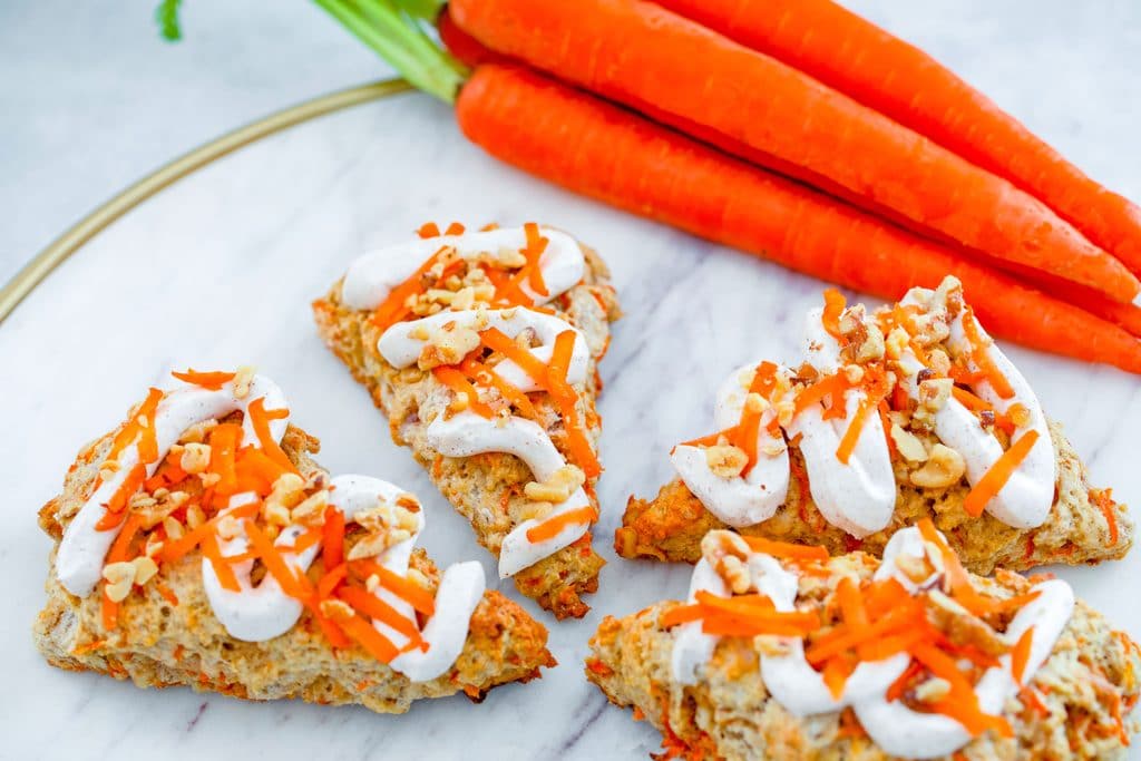 Landscape overhead view of 4 chai carrot cake scones on a marble surface, drizzled with cream cheese frosting with a bunch of carrots in the background