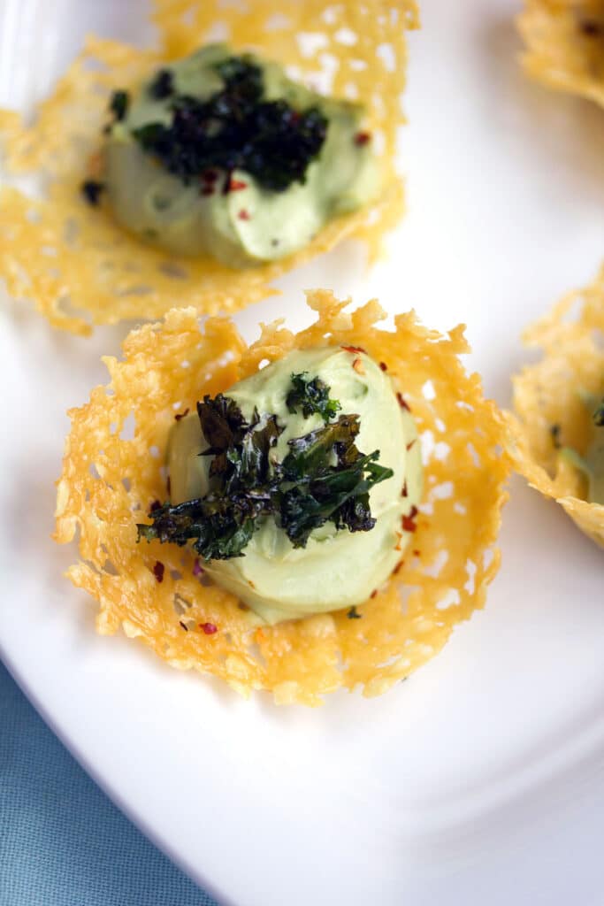 Overhead view of multiple cheddar cups filled with avocado mousse and topped with crispy kale on a white platter