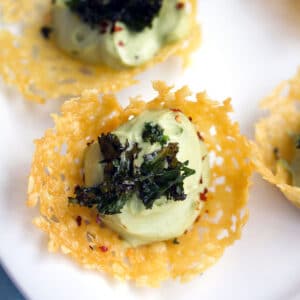 Overhead closeup view of multiple cheddar cups filled with avocado mousse and topped with crispy kale on a white platter