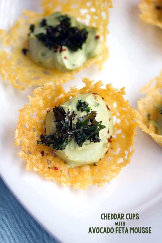 Overhead view of multiple cheddar cups filled with avocado mousse and topped with crispy kale on a white platter with recipe title at the bottom of the image