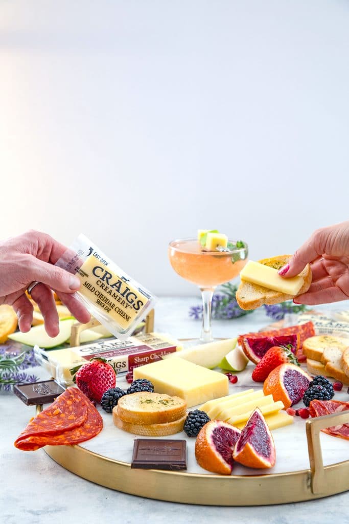 Head-on view of a cheese board with fruit, chocolate, meat, and crackers with two hands reaching out to grab cheese and a pear old Cuban cocktail in the background