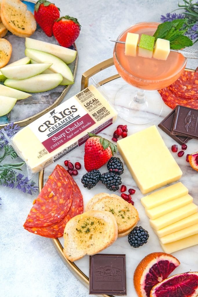 Overhead view of block of Craigs Creamery cheddar on a marble board with lots of meat, fruit, crackers, and chocolate, with pear old Cuban cocktails