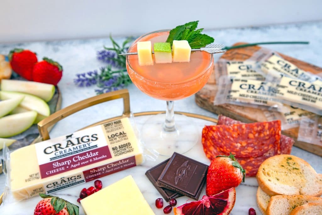 Landscape view of pear old Cuban cocktail on a marble cheeseboard featuring cheddar cheese, meats, crackers, fruits, and chocolate for a cheese and cocktail pairing party