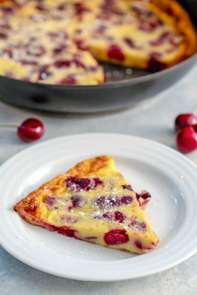 A white plate with a slice of cherry clafoutis with cherries and a skillet with whole cherry clafoutis in background