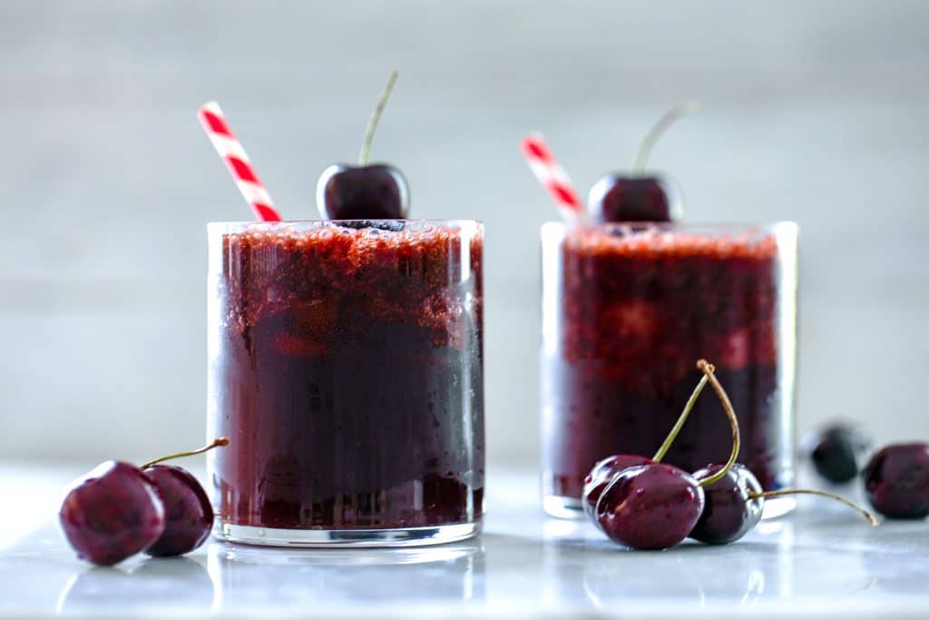 Landscape view of two glasses of cherry rum and coke with red and white striped straws, cherry garnishes, and cherries scattered on table