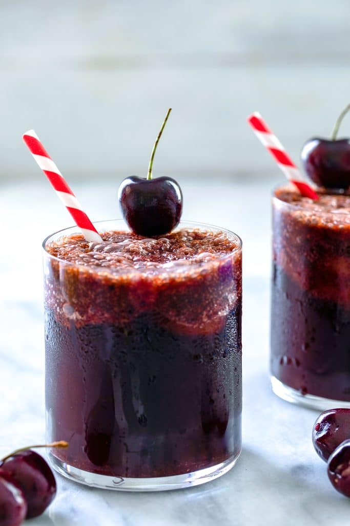 Head-on view of two glasses of cherry rum and coke with red and white striped straws, cherry garnish, and cherries on table