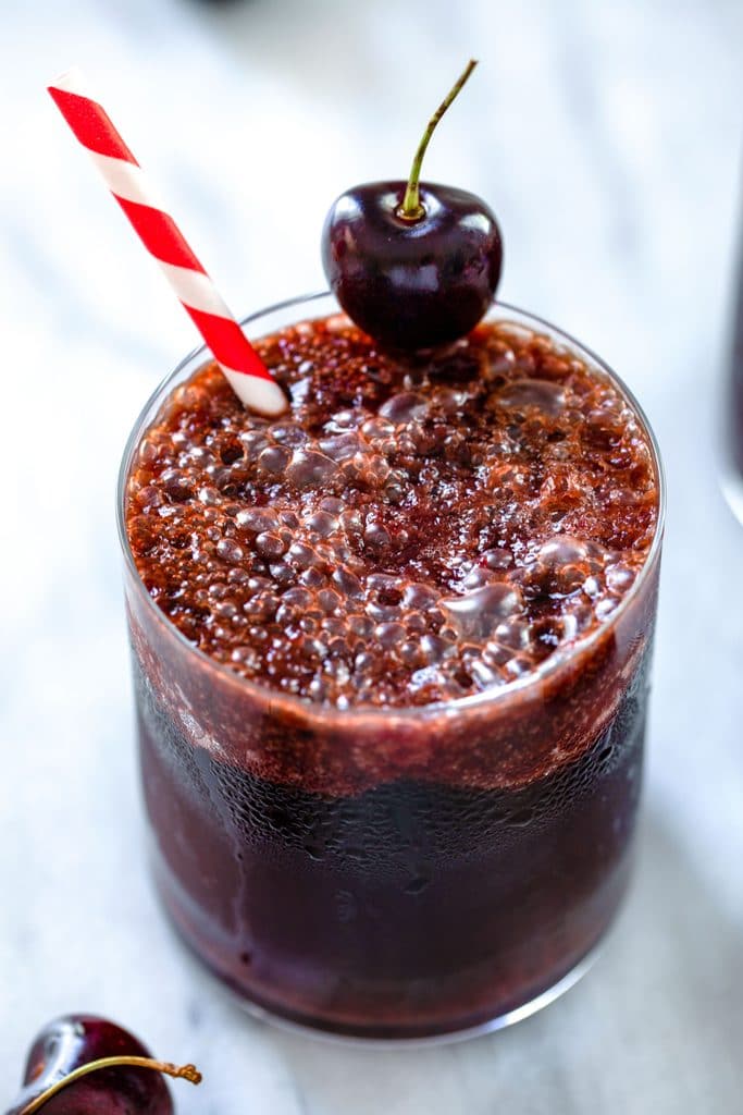 Overhead view of glass of fizzy cherry rum and coke with red and white striped straw and cherry garnish