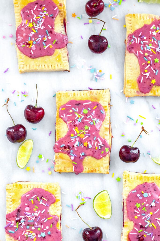 Bird's eye view of multiple cherry lime pop tarts on a marble surface with cherries, limes, and sprinkles all around