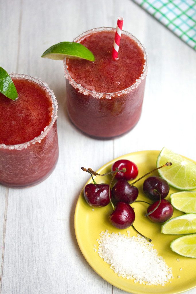 Overhead view of two cherry margaritas with lime wedge garnishes and salted rims with yellow plate with salt, cherries, and limes in foreground