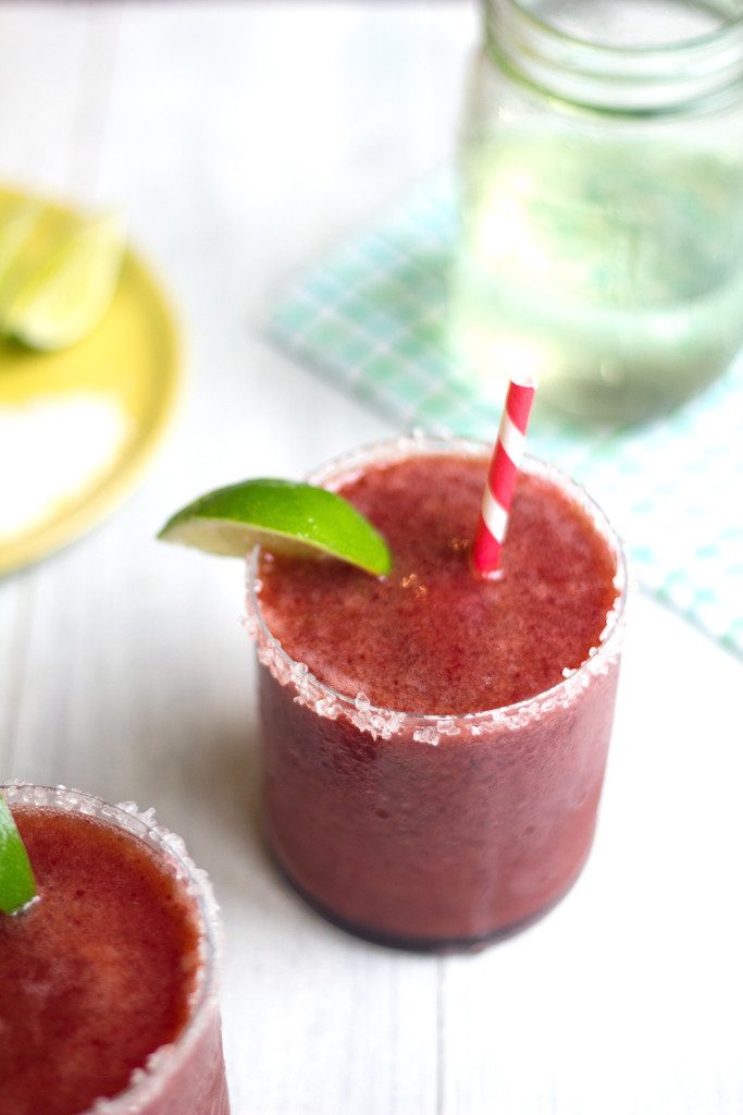 Overhead view of a cherry margarita with lime wedge garnish and salted rim with second margarita in the foreground and mason jar of simple syrup in the background