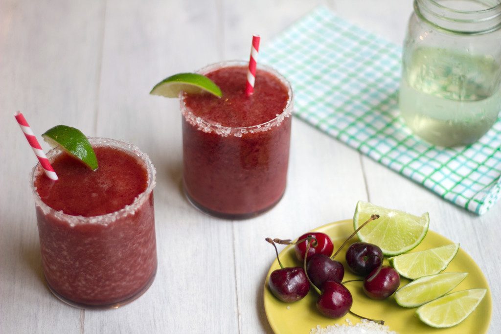 Landscape view of two cherry margaritas with lime wedge garnishes and salted rims with plate with cherries, salt, and limes in foreground and mason jar of simple syrup in background