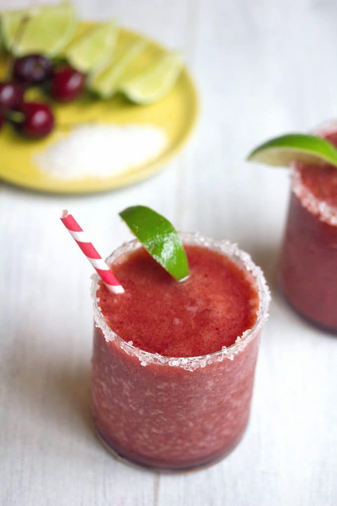 Overhead view of a cherry margarita with lime wedge garnish and salted rim with plate of salt, cherries, and lime in background