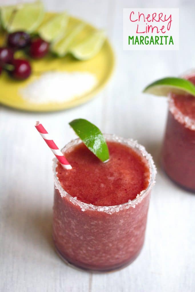 Overhead view of a cherry margarita with lime wedge garnish and salted rim with plate of salt, cherries, and lime in background and recipe title at top