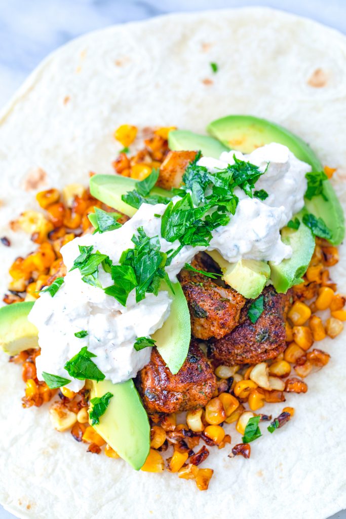 Overhead view of a flour tortilla topped with grilled corn, chicken, sliced avocado, feta sauce, and parsley