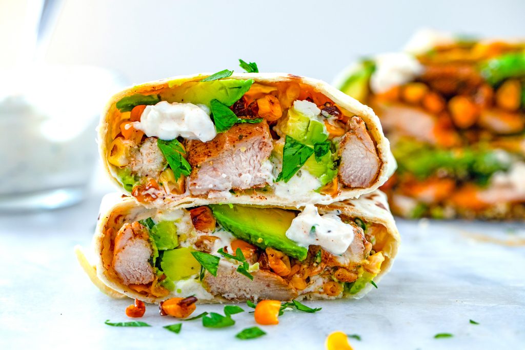 Landscape view of a chicken, avocado, and corn burrito cut in half with halves stacked on each other to showcase ingredients with feta sauce drizzled on them