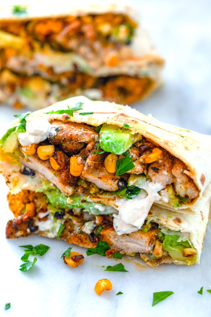 Chicken, avocado, and corn burritos cut in half and stacked on each other to showcase ingredients with feta sauce drizzled on them