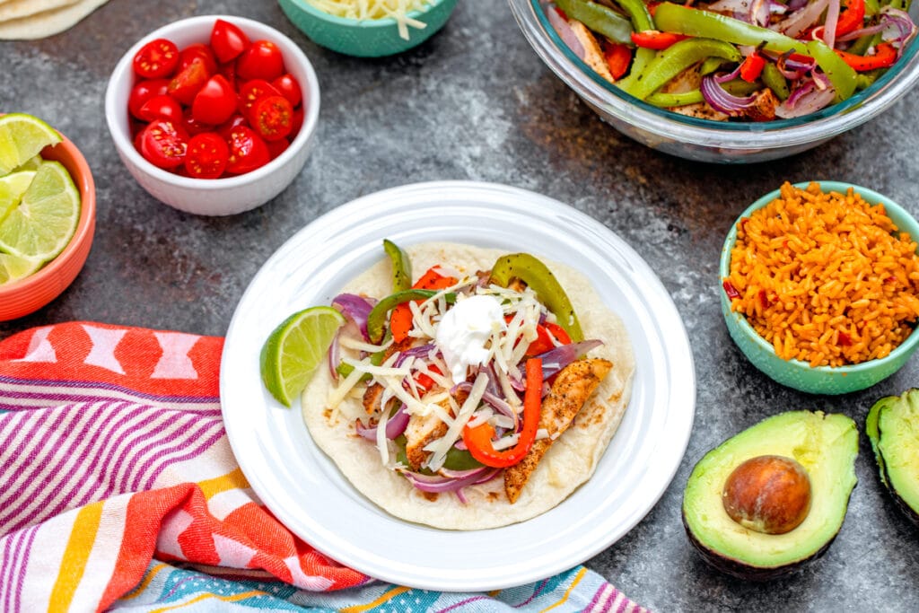 Landscape view of a chicken fajita on a plate surrounded by bowls of toppings (tomatoes, cheese, lime wedges, Spanish rice), half an avocado, and more chicken and veggies