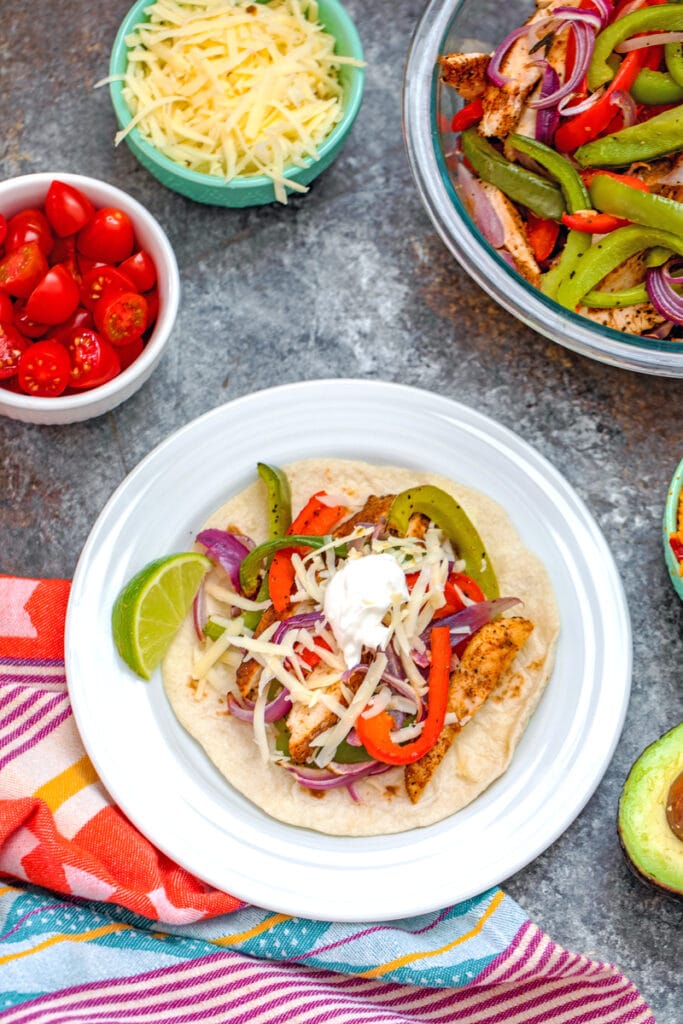 Overhead view of a prepared chicken fajita on a plate surrounded by a bowl of sliced chicken and vegetables and bowls of toppings